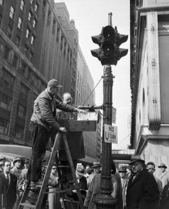 Along the lines of pedestrian signals, there were several kinds that were in use in New York City over the years. The first of its kind was introduced in Manhattan in the early 1950s. It was rather skinny, not to mention horizontal, in appearance, and its source of illumination was neon. Manufactured by the Crouse-Hinds company, this pedestrian signal was near and at Times Square, and roughly ten were in useful service for a short period of time. At the time of their introduction, they operated along with two-section (red and green) vehicular traffic signals. Below, is a photograph of an example. In the photograph, then traffic commissioner T.T. Wiley (man below the installer with hat) helps a worker install this kind of pedestrian signal at Times Square, while a group of people below them watch.